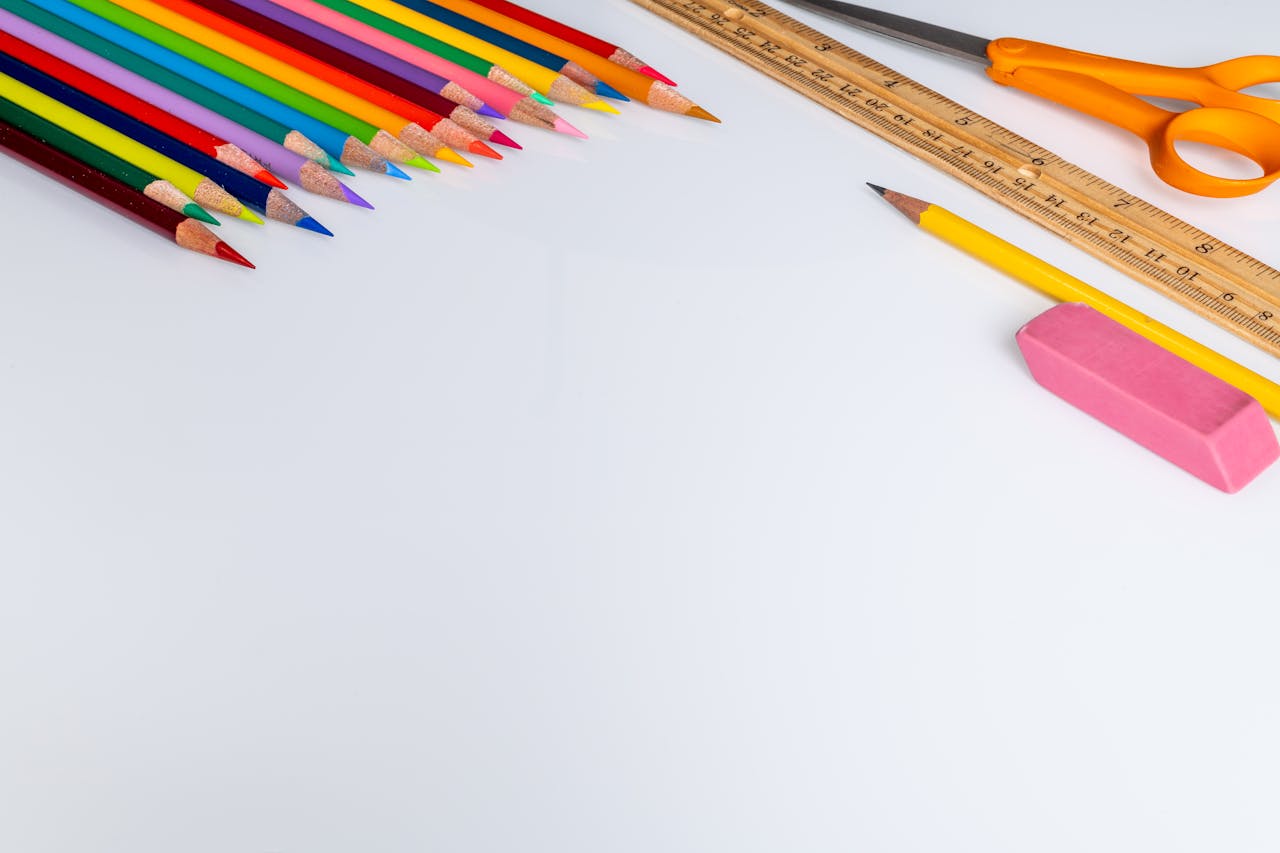 Set of colored pencils for drawing placed on white surface near wooden ruler and scissors near eraser in light studio